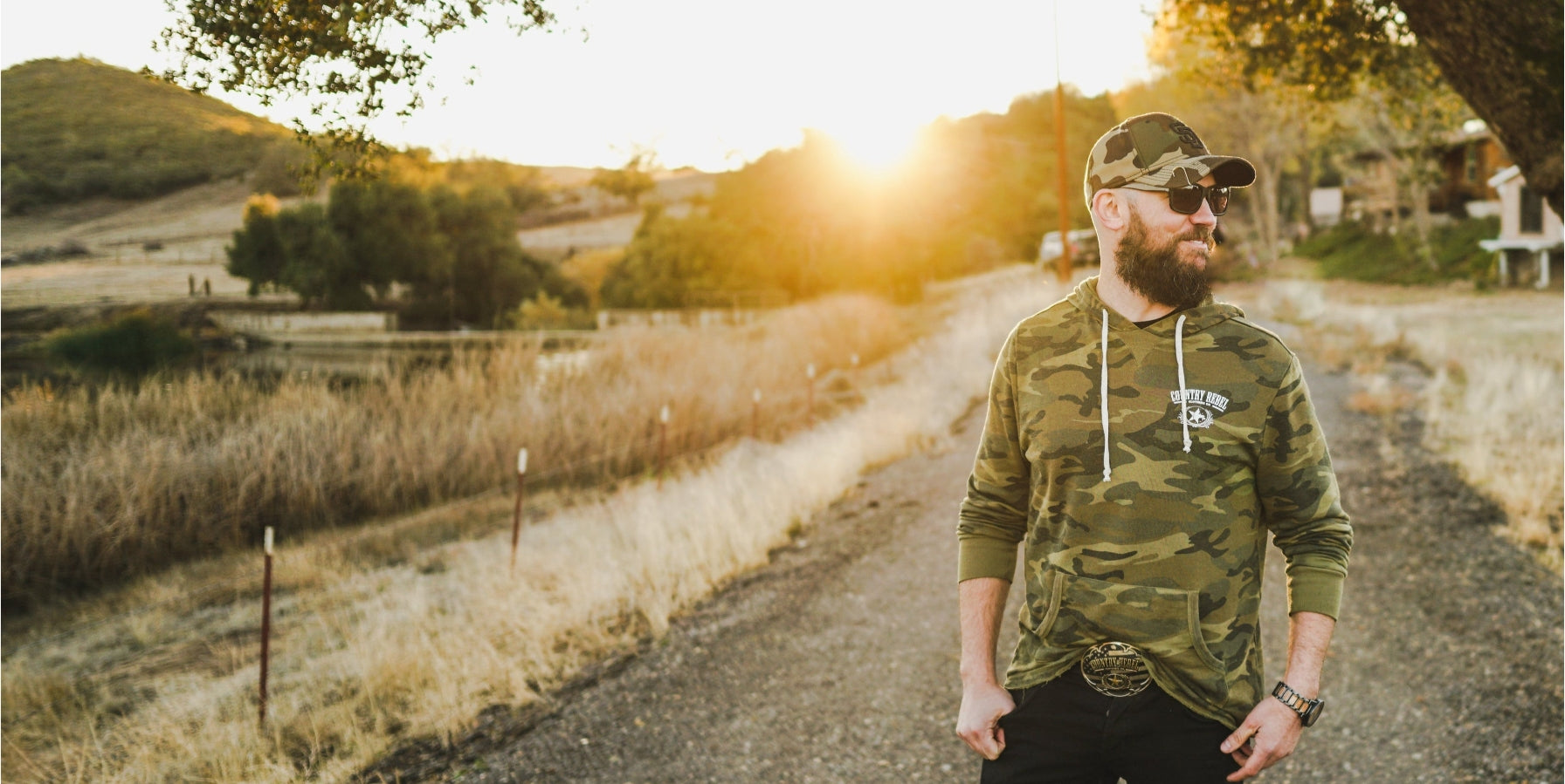 Man in camouflage shirt and cap standing on a dirt road with sunset in the background